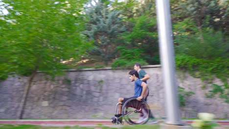 Der-Behinderte-Junge-Mann-Fährt-Schnell-Mit-Seinem-Rollstuhl-Und-Sein-Freund-Begleitet-Ihn.